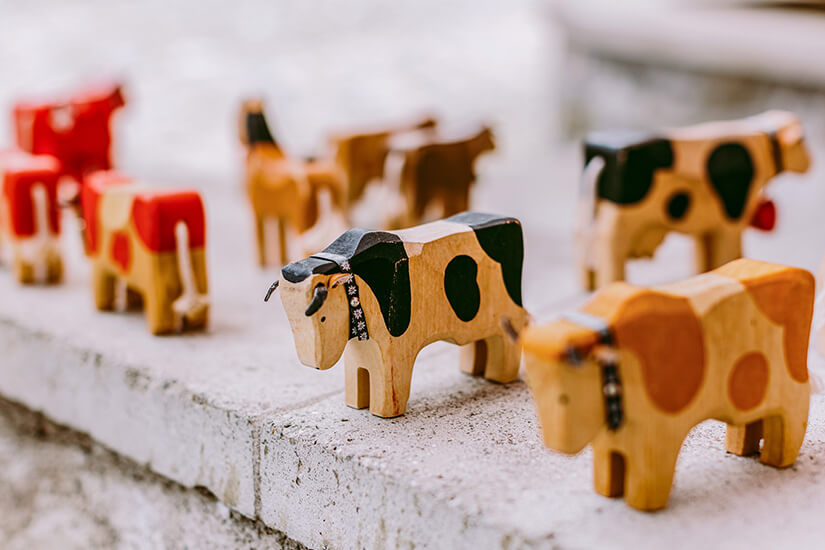 Kleine Holzfiguren in Form von Kühen stehen auf einer Steinfläche in einer Reihe. Sie sind bunt bemalt und teils mit kleinen Halsbändern verziert. Schweiz Holzspielzeug