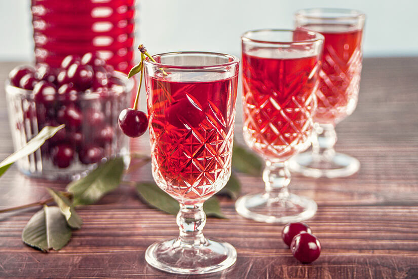 Mehrere verzierte Gläser sind mit einem roten Getränk gefüllt und auf einem Tisch arrangiert. Kirschen und Blätter liegen dekorativ daneben, einige Früchte schwimmen im Getränk. Im Hintergrund steht eine Schale mit weiteren Kirschen. Souvenir Likör