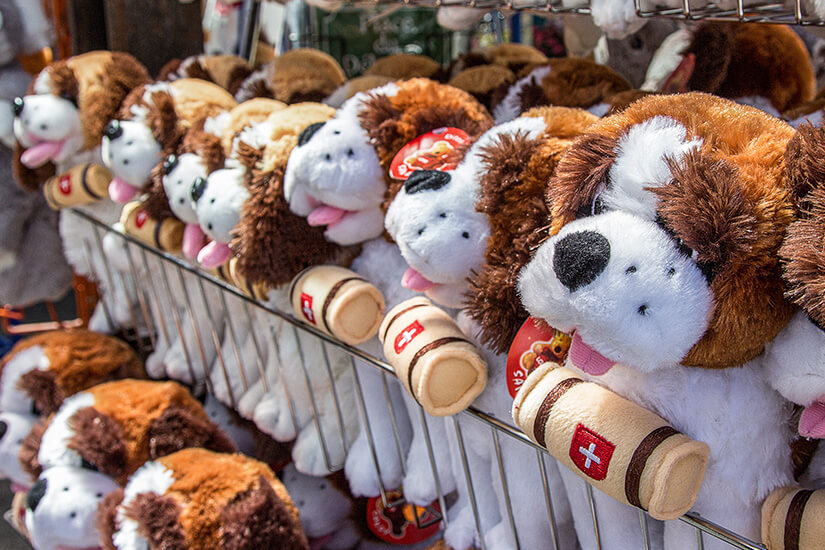 Mehrere Plüschhunde hängen dicht nebeneinander an einem Verkaufsständer. Sie haben lange Ohren, freundliche Gesichter und tragen kleine Fässchen mit Schweizer Symbolen um den Hals. Bernhadiner Kuscheltier