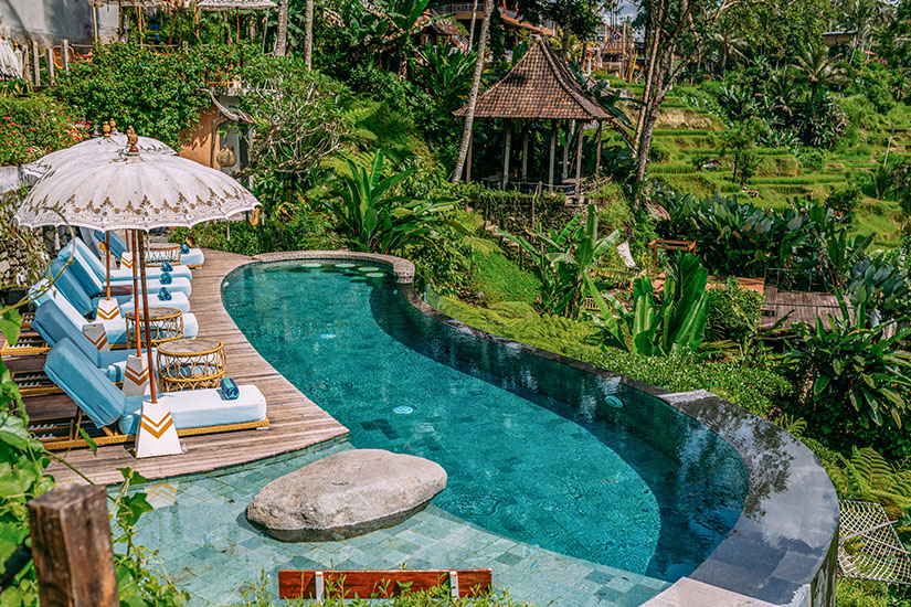 Ein geschwungener, türkisfarbener Infinity-Pool mit eleganten Sonnenliegen und einem balinesischen Sonnenschirm schmiegt sich harmonisch in eine üppige tropische Gartenlandschaft ein. Luxus Reiseziele Bali