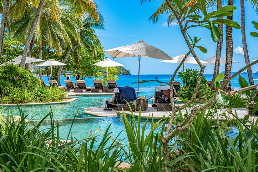 Dieser luxuriöse Hotelpool, eingebettet in üppige tropische Vegetation mit Palmen und weißen Sonnenschirmen über eleganten Liegen, öffnet sich zu einem strahlend türkisblauen Meer mit einer bewaldeten Insel im Hintergrund. Luxus Reiseziele Fidschi