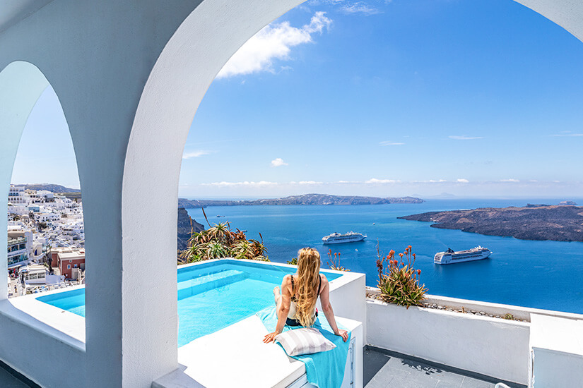 Eine junge Frau sitzt entspannt an einem kleinen Infinity-Pool auf einer weißen Terrasse und genießt den atemberaubenden Blick über die Caldera von Santorini mit ihren charakteristischen weißen Häusern und zwei Kreuzfahrtschiffen auf dem tiefblauen Ägäischen Meer. Luxus Reiseziele Santorin