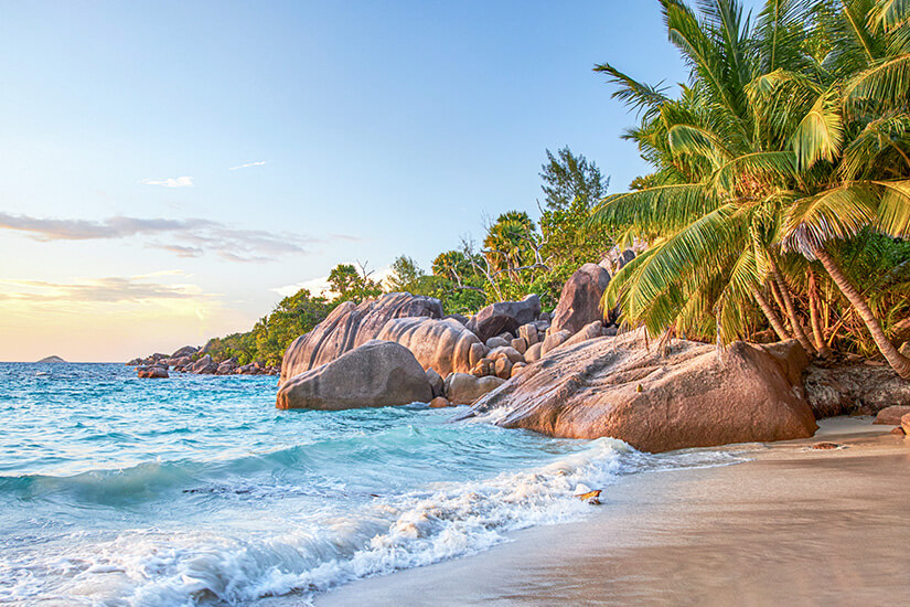 Charakteristische rötlich-graue Granitfelsen, üppige Palmen und türkisblaues Wasser mit sanft auslaufenden Wellen prägen diesen paradiesischen Strand im warmen Abendlicht. Luxus Reiseziele Seychellen