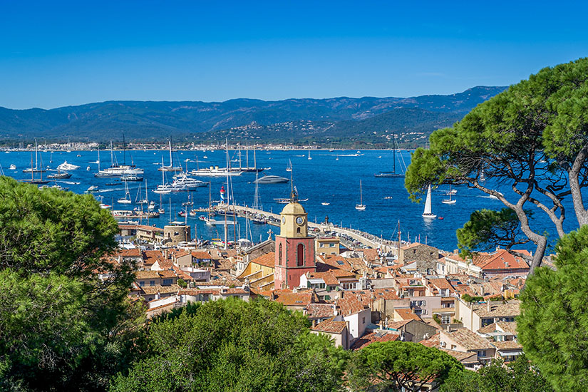 Der charakteristische Glockenturm der Kirche von Saint-Tropez überragt die terracottafarbenen Dächer der Altstadt, während der malerische Hafen mit zahlreichen Luxusyachten und Segelbooten das tiefblaue Mittelmeer vor einer bewaldeten Hügelkulisse schmückt. Frankreich Saint Tropez