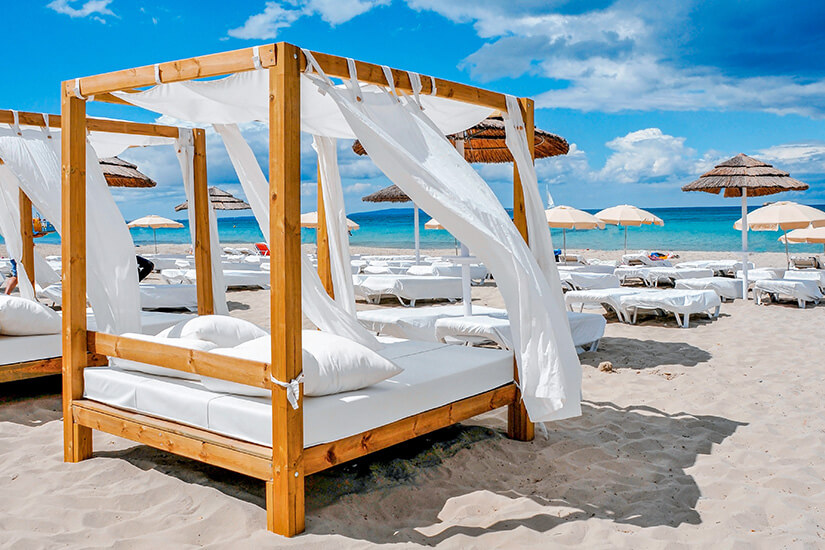 Ein elegantes Holz-Himmelbett mit wehenden weißen Vorhängen dominiert den Vordergrund eines stilvollen Beachclubs, während sich dahinter Reihen weißer Sonnenliegen unter Strohschirmen bis zum türkisfarbenen Meer erstrecken. Luxus Reiseziele Ibiza