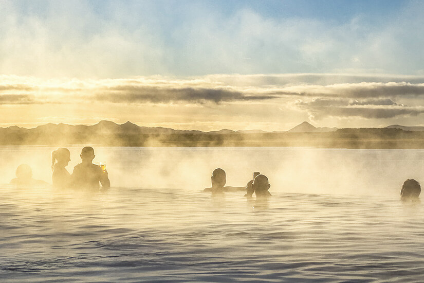 Silhouetten mehrerer Menschen tauchen in einem dampfenden geothermalen Naturbad, während die tiefstehende Sonne den aufsteigenden Wasserdampf in goldenes Licht taucht und die Bergkulisse im Hintergrund mystisch erscheinen lässt. Luxus Reiseziele Island