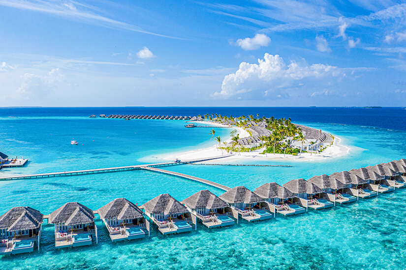 Eine atemberaubende Luftaufnahme zeigt Reihen von Overwater-Bungalows mit Strohdächern, die sich auf langen Stegen über das strahlend türkisfarbene Wasser eines Atolls erstrecken und eine kleine, palmenbestandene Insel mit weißem Sandstrand umrahmen. Luxus Reiseziele Malediven