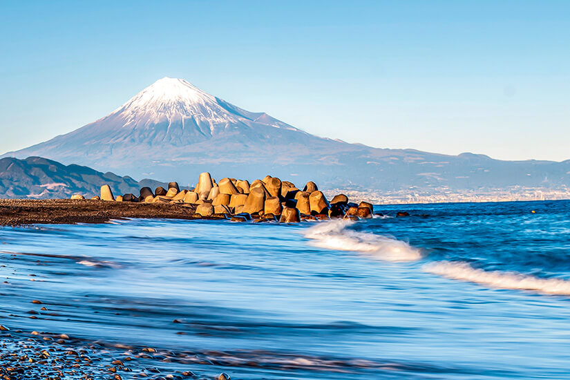 Sanfte Wellen rollen an einen Kiesstrand, neben dem große Wellenbrecher aus Beton ins Meer ragen. Im Hintergrund erhebt sich ein markanter, schneebedeckter Vulkan vor klarem Himmel. Japan Miho no Matsubara