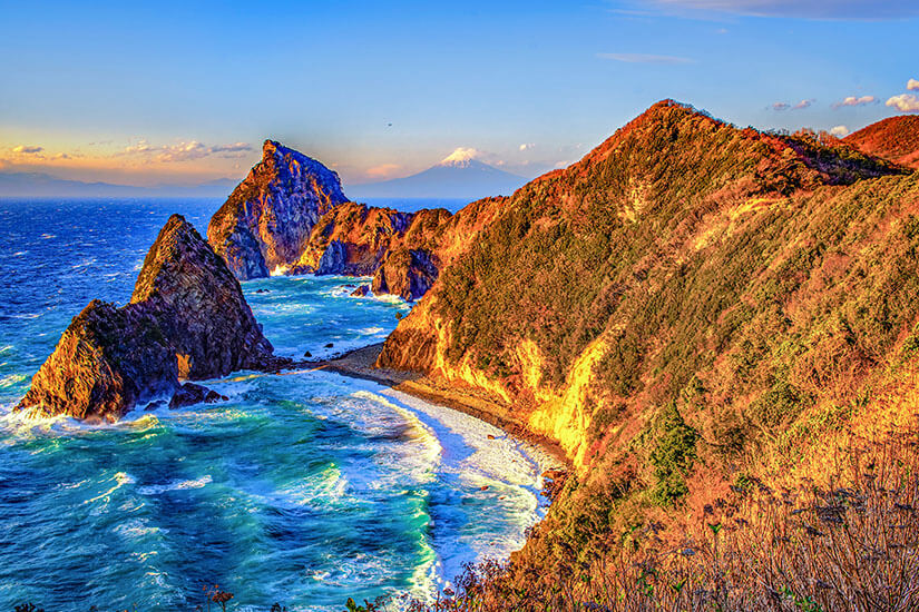 Raues Meer trifft auf steile, zerklüftete Felsen entlang einer dramatischen Küste. Die Hänge leuchten im warmen Licht, während im Hintergrund ein schneebedeckter Berg am Horizont sichtbar ist. Wellen schlagen gegen die Felsen. Japan Izu