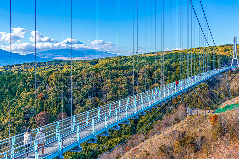 Eine lange Hängebrücke spannt sich über ein bewaldetes Tal, auf der mehrere Menschen unterwegs sind. In der Ferne erhebt sich ein markanter, schneebedeckter Berg über die Landschaft. Japan Skywalk