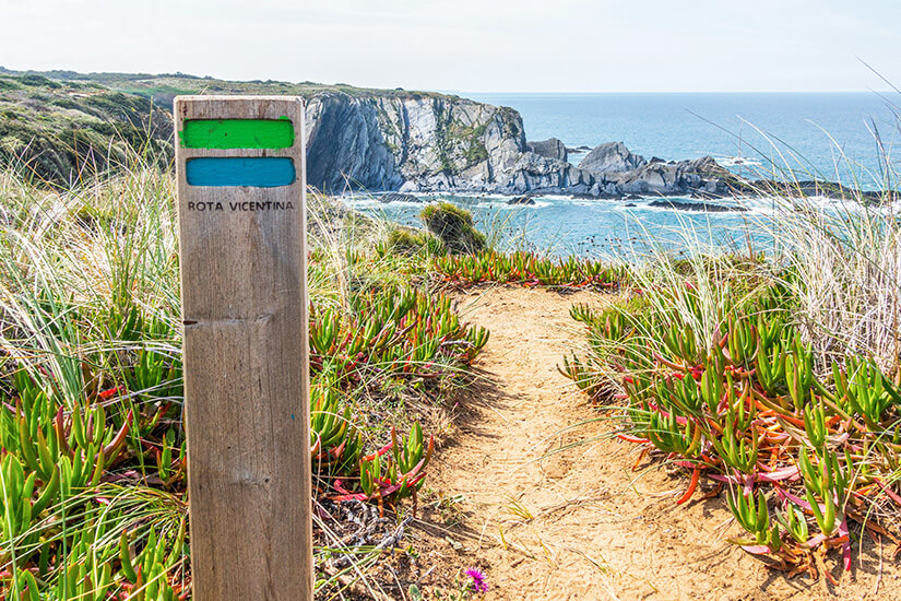 Eine Holzmarkierung mit der Aufschrift "Rota Vicentina" steckt neben einem sandigen Pfad, der durch bunte Vegetation führt. Im Hintergrund sind Klippen und blaues Meer zu sehen. Fischerweg Portugal Willkommen