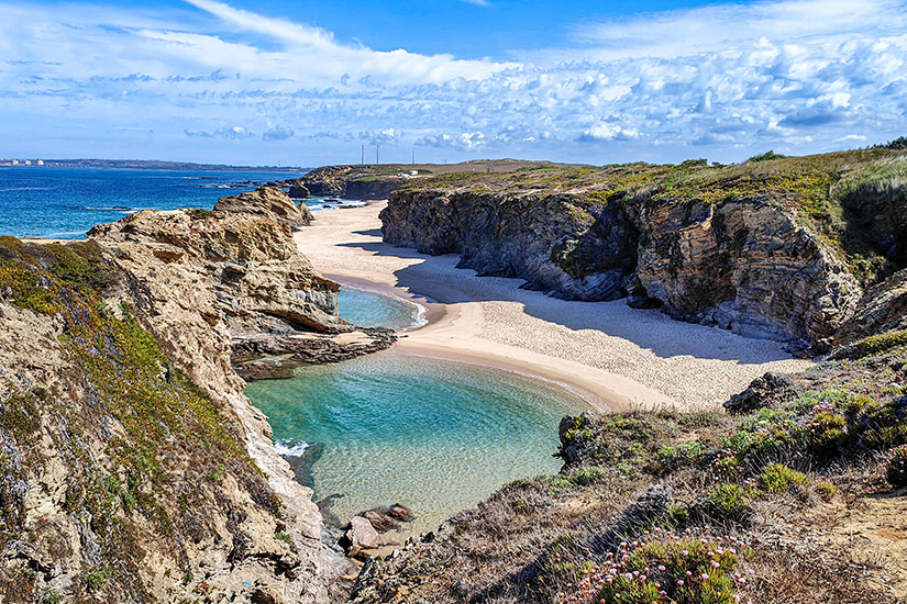 Ein heller Sandstrand liegt vor eindrucksvollen grünen Klippen. Das Meerwasser leuchtet in klaren Türkistönen. Fischerweg Portugal Strand