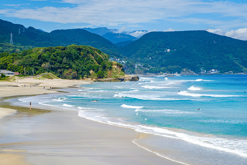 Ein weiter Sandstrand zieht sich entlang einer geschwungenen Küste mit sanften Wellen. Einige Menschen befinden sich im Wasser oder spazieren am Ufer, während grüne Hügel die Landschaft im Hintergrund prägen. Shizuoka Strand