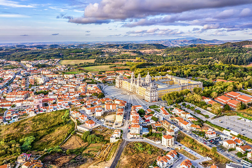 Eine weitläufige Stadtlandschaft breitet sich aus, durchzogen von Straßen und dicht bebauten Häusern mit roten Dächern. Im Zentrum steht ein großes, beeindruckendes Schloss- oder Klostergebäude mit symmetrischer Architektur. Umgeben ist alles von grünen Feldern und sanften Hügeln unter einem teils bewölkten Himmel. Mafra Stadt