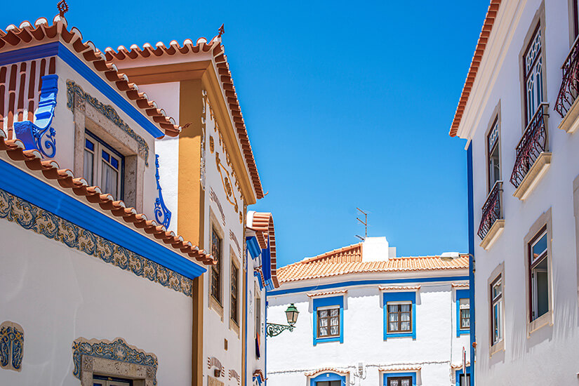 Enge Gasse mit hell verputzten Häusern, die mit blauen Zierelementen und Fliesen geschmückt sind. Rote Ziegeldächer und kleine Balkone verleihen der Architektur einen mediterranen Charakter. Über den Gebäuden spannt sich ein klarer, tiefblauer Himmel. Portugal Ericeira