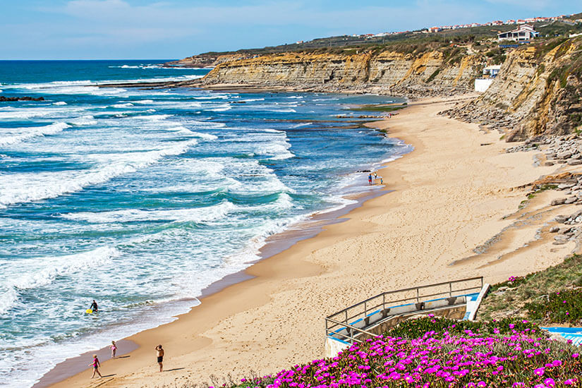 Ein weiter Sandstrand erstreckt sich entlang einer felsigen Küste mit hohen Klippen. Wellen rollen auf das Ufer zu, während einige Menschen am Strand spazieren oder im Wasser sind. Im Vordergrund blühen farbenfrohe Pflanzen und ein kleiner Weg führt hinunter zum Strand. Portugal Surfen