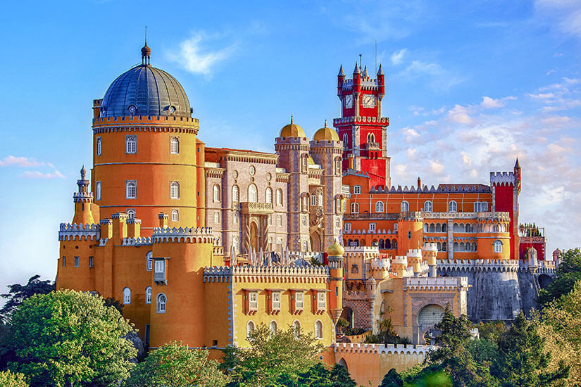 Ein farbenprächtiges, märchenhaft wirkendes Schloss mit Türmen, Kuppeln und verspielten Details erhebt sich über einer grünen Landschaft. Die Architektur kombiniert verschiedene Stile und leuchtet in Gelb-, Rot- und Weißtönen. Der Himmel darüber ist leicht bewölkt. Portugal Sintra