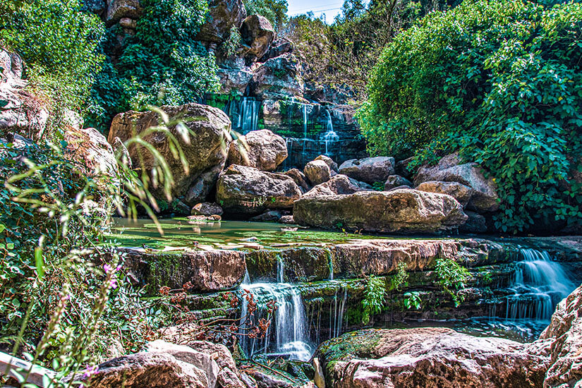 Ein kleiner Wasserfall fließt über mehrere Felsstufen in ein ruhiges Becken. Große Steine und dichtes Grün umgeben die Szenerie und verleihen ihr eine natürliche, abgeschiedene Atmosphäre. Portugal Wasserfall