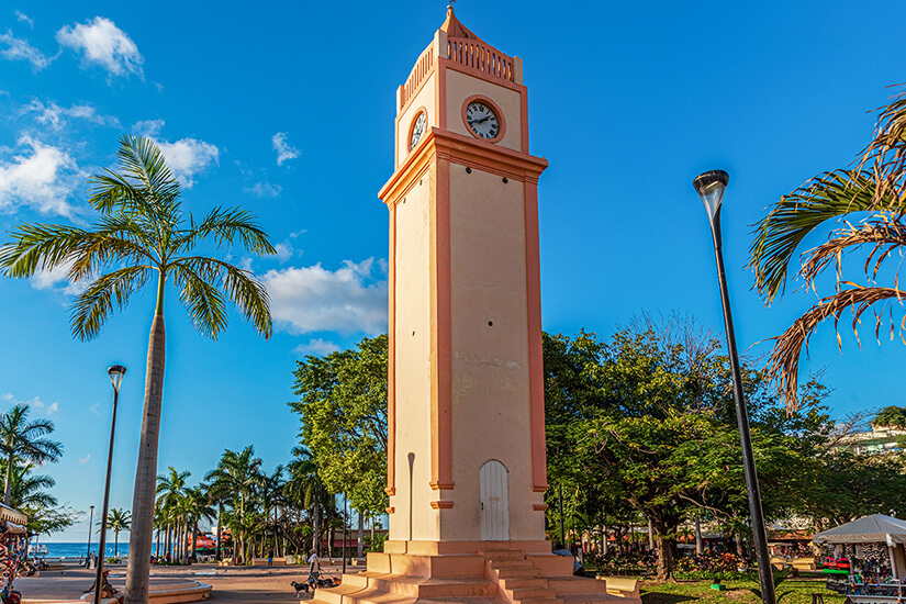 Ein hoher, beigefarbener Uhrturm mit lachsfarbenen Akzenten steht zentral auf einem sonnigen Platz in San Miguel de Cozumel unter blauem Himmel mit Palmen. Cozumel Mexiko San Miguel