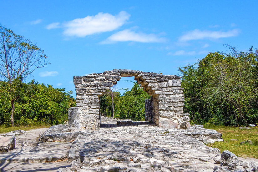 Ein steinerner Torbogen einer Maya-Ruine aus grauen Steinblöcken steht auf einem felsigen Plateau, umgeben von dichtem grünem Gebüsch unter einem strahlend blauen Himmel. Mexiko San Gervasio Cozumel Mexiko San Gervasio