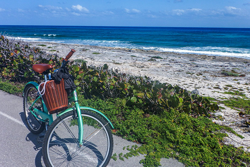 Ein türkisfarbenes Fahrrad mit einem braunen Weidenkorb steht auf einem asphaltierten Weg neben grüner Vegetation, mit Blick auf eine felsige Küste und das blaue Meer unter einem klaren Himmel auf Cozumel. Cozumel Mexiko Radfahren