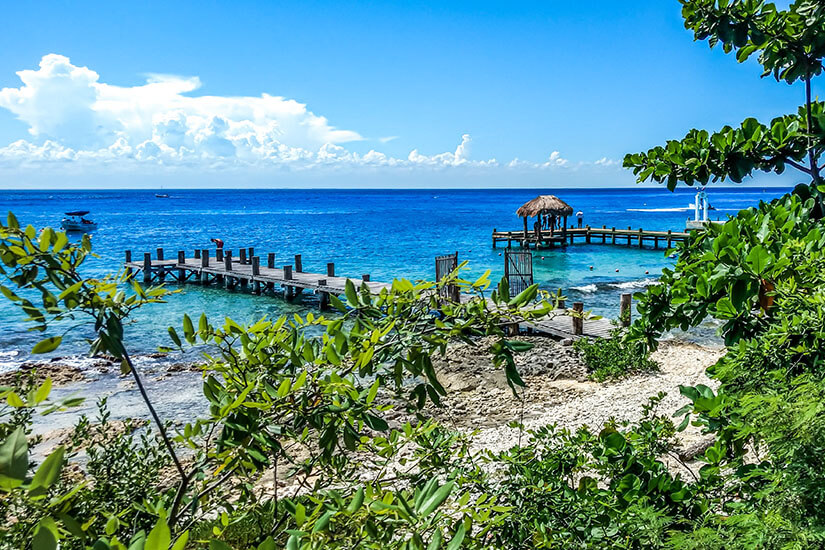 Blick durch üppiges grünes Laub auf zwei rustikale Holzstege, die in kristallklares, türkisfarbenes Wasser führen. Am Ende des einen Stegs steht eine kleine Hütte mit Palmendach. Der blaue Himmel ist leicht bewölkt und das Meer ruhig. Cozumel Mexiko Bucht