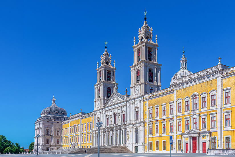 Ein großes, barockes Gebäude mit gelb-weißer Fassade und zwei hohen Türmen dominiert die Szene. Die Architektur ist reich verziert mit Säulen, Kuppeln und symmetrischen Elementen. Vor dem Bauwerk liegt ein weiter Platz unter klarem, blauem Himmel. Mafra Palast