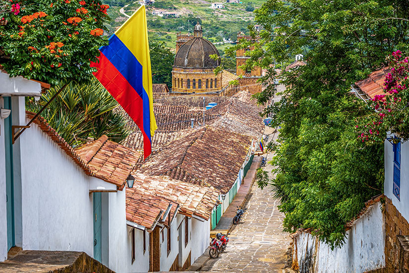 Eine kolumbianische Flagge weht stolz über den terracottafarbenen Ziegeldächern einer malerischen Kolonialstadt, deren kopfgepflasterte Gasse hinunter zu einer imposanten Kirchenkuppel führt. Kolumbianisches Essen Barichara