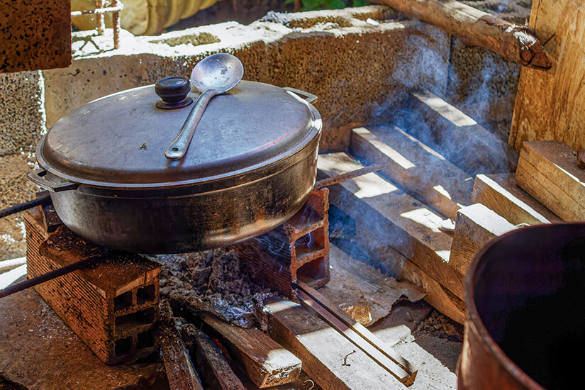 Ein schwerer gusseiserner Topf mit Kelle dampft auf einem improvisierten Ziegelsteinherd über offenem Holzfeuer, während Rauch durch den rustikalen Unterstand zieht. Kolumbianisches Essen Kochstelle