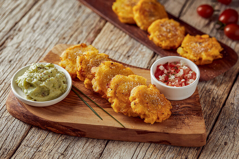 Auf einem rustikalen Holzbrett sind knusprig frittierte Kochbananenscheiben angerichtet, begleitet von zwei Dipsaucen – einer cremigen Guacamole und einer frischen Tomatensalsa. Kolumbianisches Essen Patacones