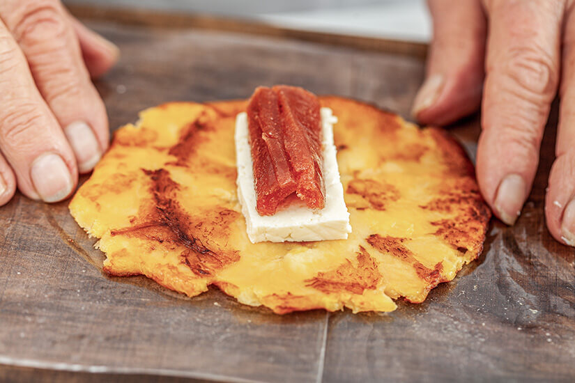 Zwei Hände halten eine flach gedrückte, goldbraun gebratene Kochbanane, auf der ein Stück weißer Frischkäse und ein Streifen rötlicher Guavenpaste angerichtet sind. Kolumbianisches Essen Aborrajados
