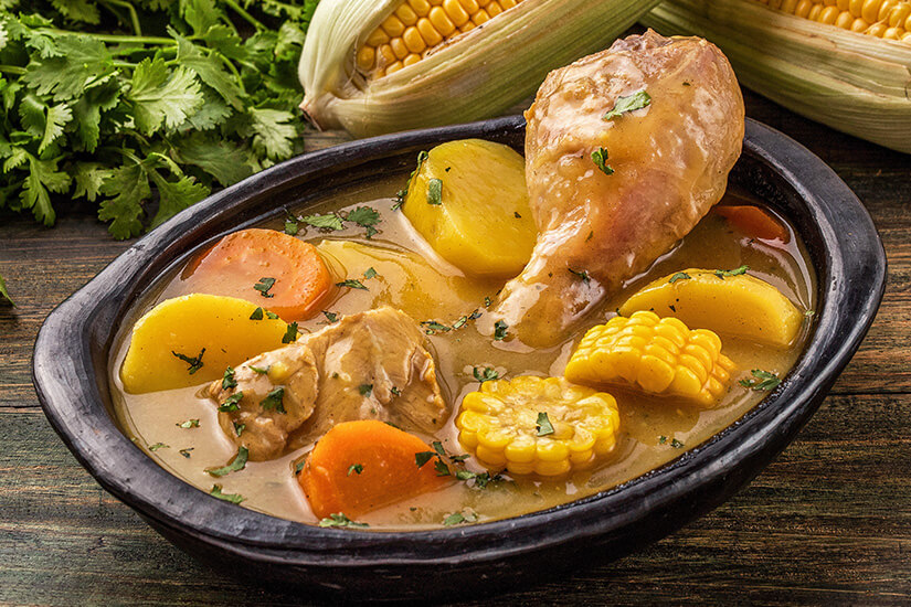 Eine reichhaltige Hühnersuppe mit Kartoffeln, Karotten, Maiskolben und Koriander dampft in einem rustikalen schwarzen Tongefäß, während frische Zutaten im Hintergrund die Natürlichkeit des Gerichts unterstreichen. Kolumbianisches Essen Sancocho