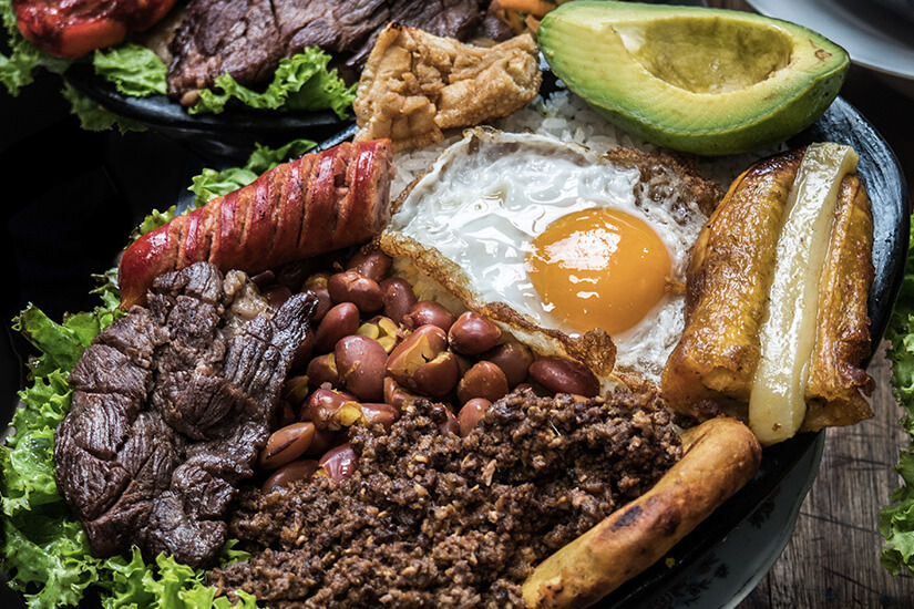 Das kolumbianische Nationalgericht schlechthin, präsentiert auf einem Teller eine beeindruckende Vielfalt an Komponenten: gegrilltes Fleisch, Hackfleisch, rote Bohnen, Spiegelei, Avocado, gebratene Kochbanane, Chicharrón und Reis. Kolumbianisches Essen Bandeja Paisa
