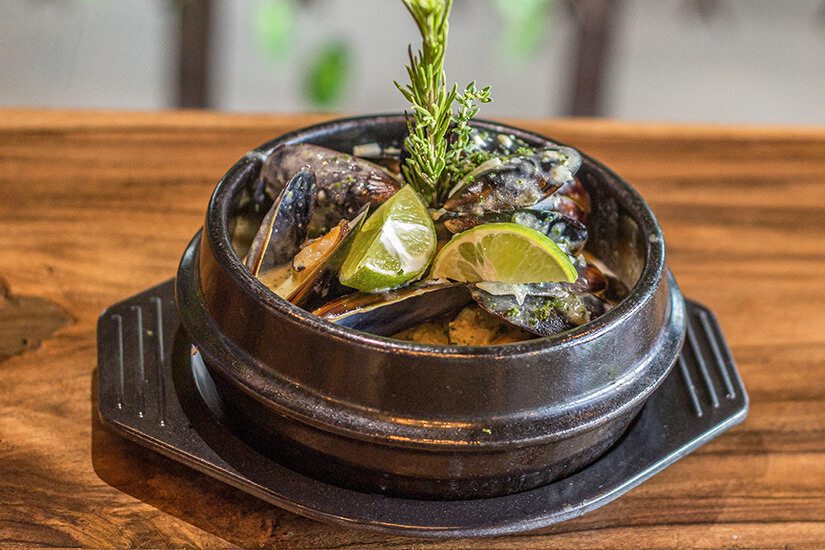 Gedämpfte Miesmuscheln in einem schwarzen gusseisernen Topf, verfeinert mit frischen Kräutern und Limettenspalten, werden auf einem rustikalen Holzuntersetzer serviert und verströmen das Flair eines gehobenen Meeresfrüchterestaurants. Cazuela de Mariscos
