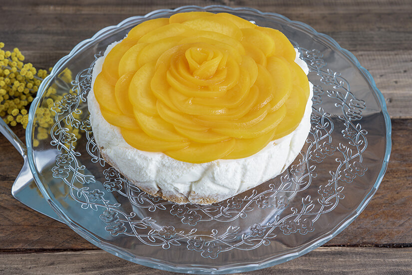 Eine elegante Torte auf einem verzierten Glasteller besticht durch ihren kunstvoll zur Rosenform arrangierten goldgelben Pfirsichbelag. Kolumbianisches Essen Tres Leches