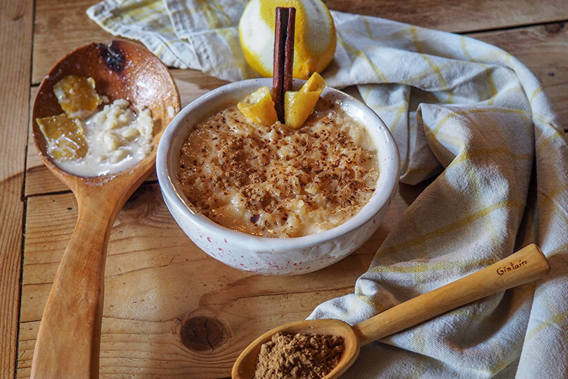Eine cremige Reismilch in einer weißen Schüssel ist mit gemahlenem Zimt bestreut und mit frischen Zitronenstücken garniert, während Holzlöffel und ein Leinentuch das rustikale Ambiente unterstreichen. Kolumbianisches Essen Milchreis