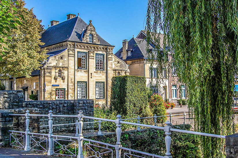 Eine Brücke führt über ein Gewässer umgeben von viel Grün. Im Hintergrund sind Residenzhäuser zu sehen. Valkenburg Niederlande Willkommen