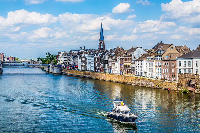 Der Fluss Maas führt durch die Stadt Maastricht. Weiße Häuserfassaden, eine Brücke und ein Boot sind zu sehen. Valkenburg Niederlande Maastricht