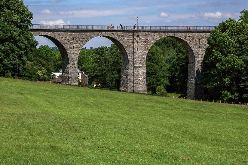 Eine Steinbrücke mit hohen Bögen führt über eine grüne Wiese und Bäume. Radfahrer sind auf der Brücke zu sehen. Valkenburg Niederlande Radfahren