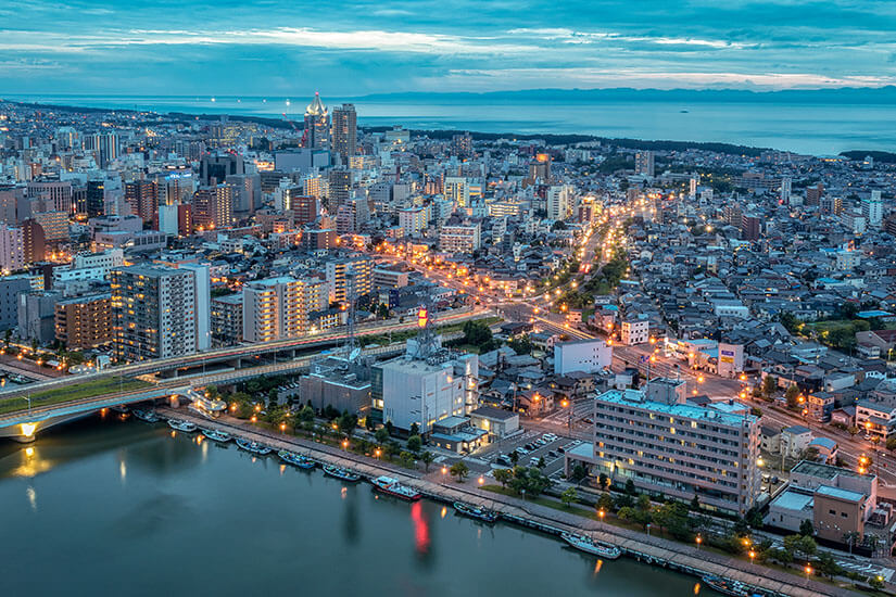 Luftaufnahme von Niigata-Stadt bei blauer Stunde und Dämmerung, mit beleuchteten Straßen, Gebäuden, einer Brücke über den Shinano-Fluss, der ins Meer mündet, und Bergen am Horizont unter klarem Himmel. Niigata Japan Vogelperspektive