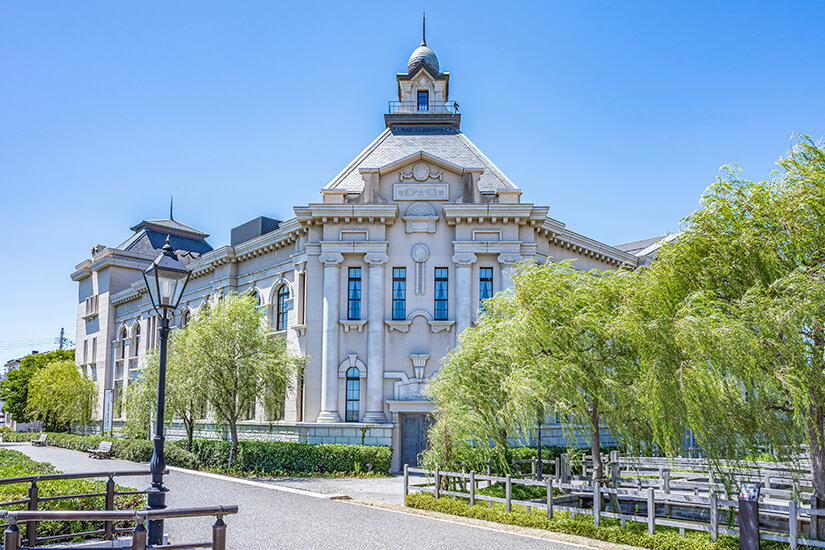Das historische Museum zur Stadtgeschichte in Niigata ist ein prachtvolles, helles Gebäude im westlichen Architekturstil mit imposanten Säulen und einem markanten Turmdach, umgeben von grünen Trauerweiden unter blauem Himmel. Niigata Japan Minatopia