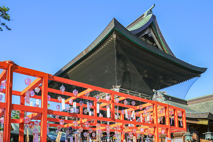 Zahlreiche kleine, gläserne Windglocken hängen an einem leuchtend orangefarbenen Holzgerüst vor dem imposanten, dunklen Holzdach des Hakusan-Schreins in Niigata unter blauem Himmel. Niigata Japan Hakusan Schrein