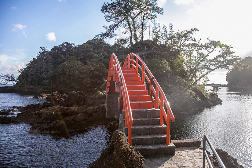 Eine steile, leuchtend orangefarbene Brücke mit Stufen führt über dunkle Felsen im Meer zu einer kleinen, dicht bewaldeten Insel unter einem hellen, leicht bewölkten Himmel. Niigata Japan Insel Sado