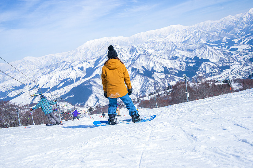 Eine Snowboarderin in einer senfgelben Jacke blickt von einer verschneiten Piste auf ein beeindruckendes Panorama aus schneebedeckten Berggipfeln unter einem hellblauen Himmel. Niigata Japan Skifahren