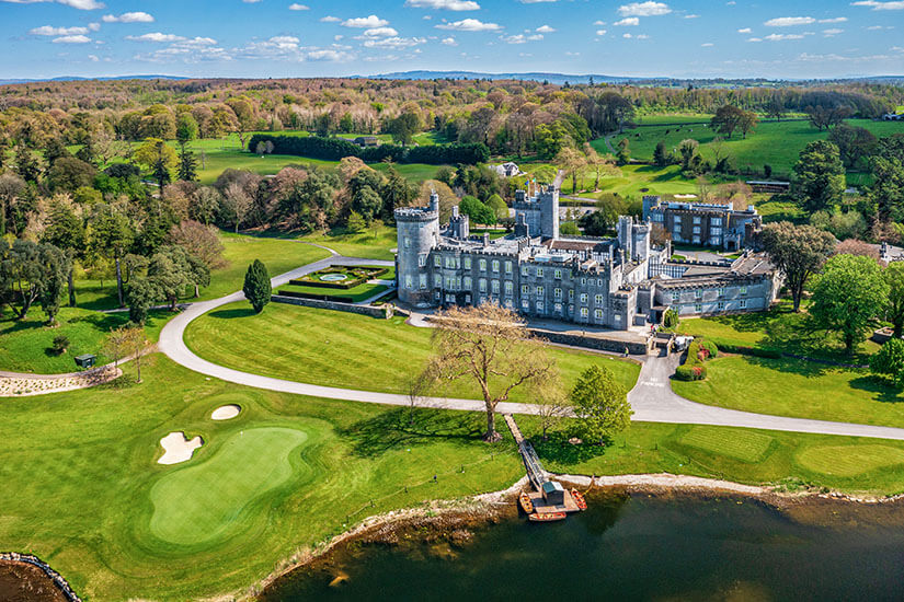 Ein imposantes Schlosshotel thront inmitten saftig grüner Parklandschaft direkt am Seeufer. Davor erstreckt sich ein gepflegtes Green mit weißem Bunker, umgeben von altem Baumbestand. Die Szenerie verbindet Geschichte, Natur und Golf zu einem Bild, das typischer für die Grüne Insel kaum sein könnte. Golfdestinationen weltweit Irland
