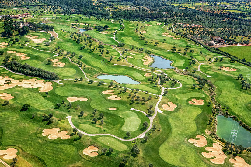 Aus der Vogelperspektive erstreckt sich eine weitläufige Golfanlage mit zahlreichen Bunkern, mehreren Wasserhindernissen und von Olivenbäumen gesäumten Fairways. Die mediterrane Landschaft leuchtet in warmen Grün- und Sandtönen. Ein Platz, der strategisches Spiel auf höchstem Niveau verlangt. Golfdestinationen weltweit Spanien