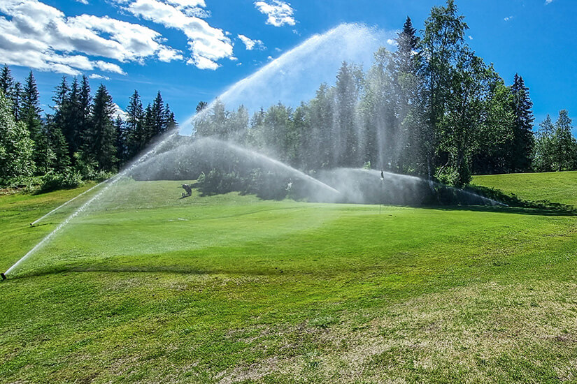 Sprinkler bewässern ein weitläufiges Green, eingerahmt von dichten Nadelbäumen unter strahlend blauem Sommerhimmel. Die Weite der Landschaft und das satte Grün vermitteln die besondere Ruhe skandinavischer Golfplätze. Hier spielt man fernab vom Trubel, umgeben von unberührter Natur. Golfdestinationen weltweit Norwegen