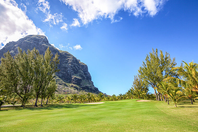 Ein sattgrünes Fairway zieht sich durch tropische Vegetation, während im Hintergrund ein gewaltiger Felsgipfel steil in den Himmel ragt. Palmen säumen den Kurs, darüber spannt sich ein leuchtend blauer Himmel mit wenigen Wolken. Hier verschmelzen Strandparadies und Championship-Golf zu einem einzigartigen Erlebnis. Golfdestinationen weltweit Mauritius