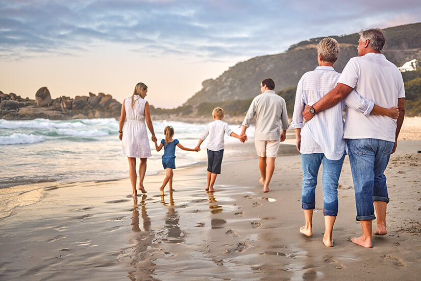 Eine Mehrgenerationenfamilie in hellen, sommerlichen Outfits spaziert Hand-in-Hand am Strand entlang, während die Wellen sanft ans Ufer rollen und die Abendsonne die Szenerie in warmes Licht taucht. Packliste Sommerurlaub Familie