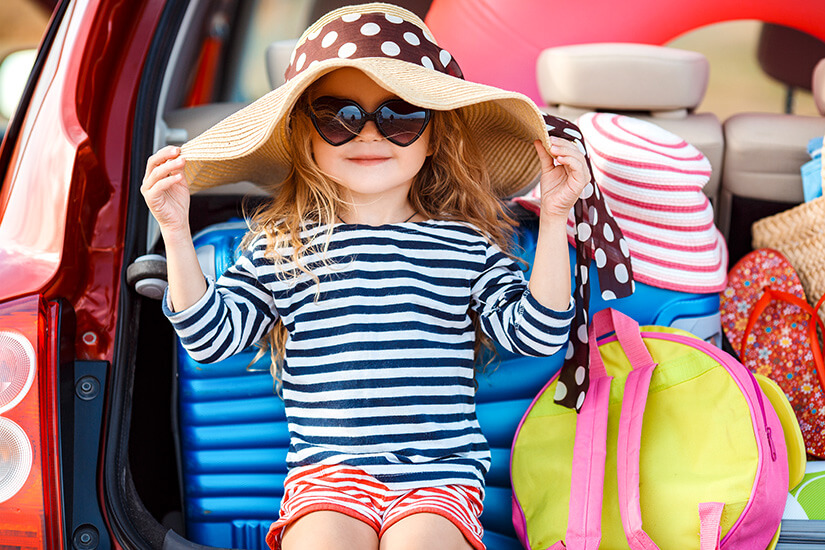 Ein charmantes kleines Mädchen mit herzförmiger Sonnenbrille und einem großen Strohhut sitzt lässig im geöffneten Kofferraum eines Autos, umgeben von buntem Gepäck, Rucksäcken und Flipflops. Packliste Sommerurlaub Kleinkind